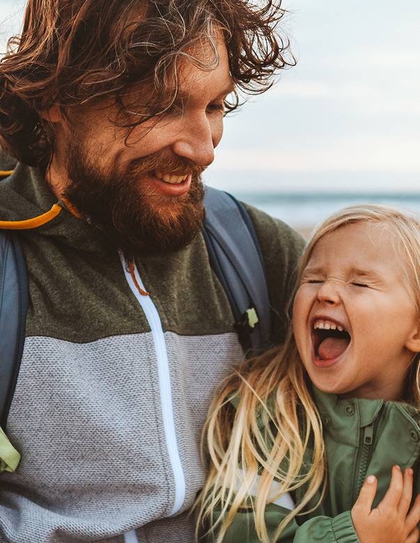 Bearded man with long hair holding blond child on lap during hike.