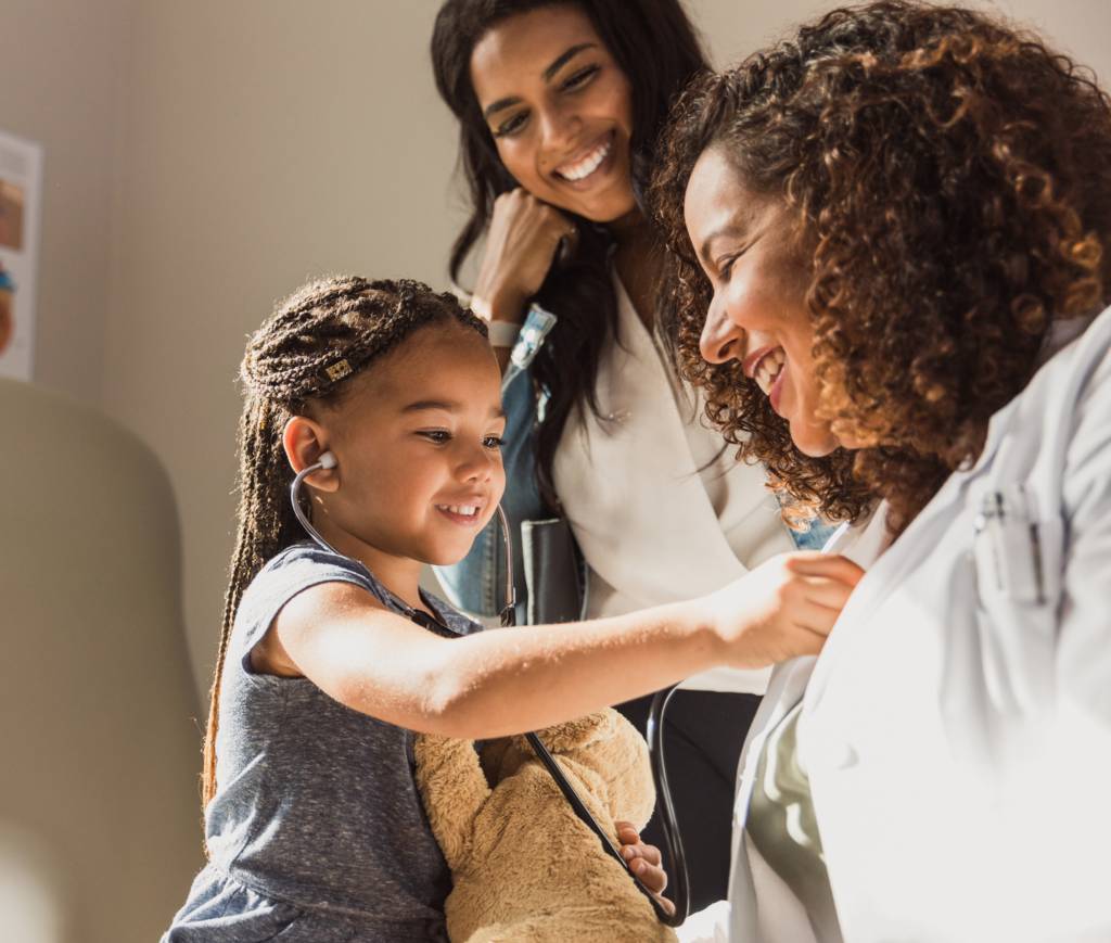 Young child at doctors office holding teddy bear, using stethoscope on doctor while mom looks on.