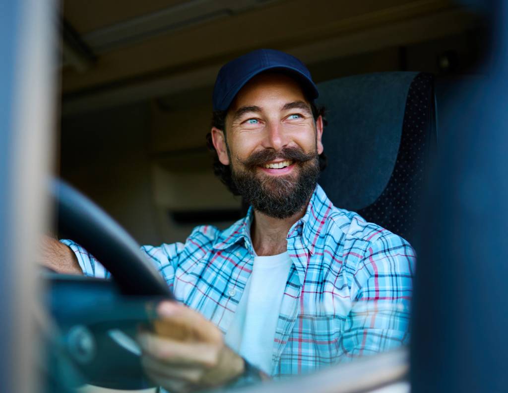 Bearded man in blue hat sits in drivers seat of large truck.
