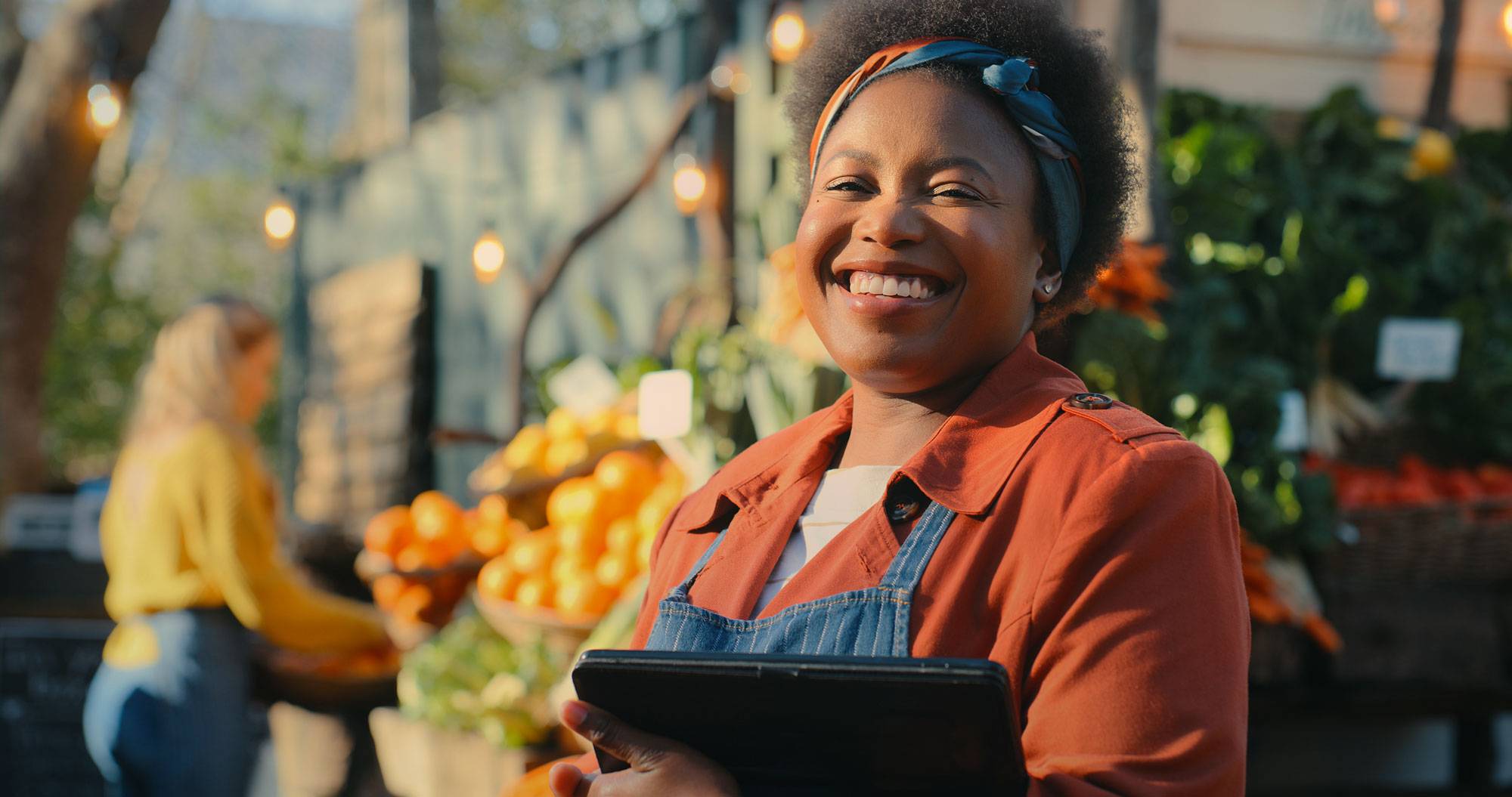 Woman in apron in front of farm stand, smiling and holding tablet.