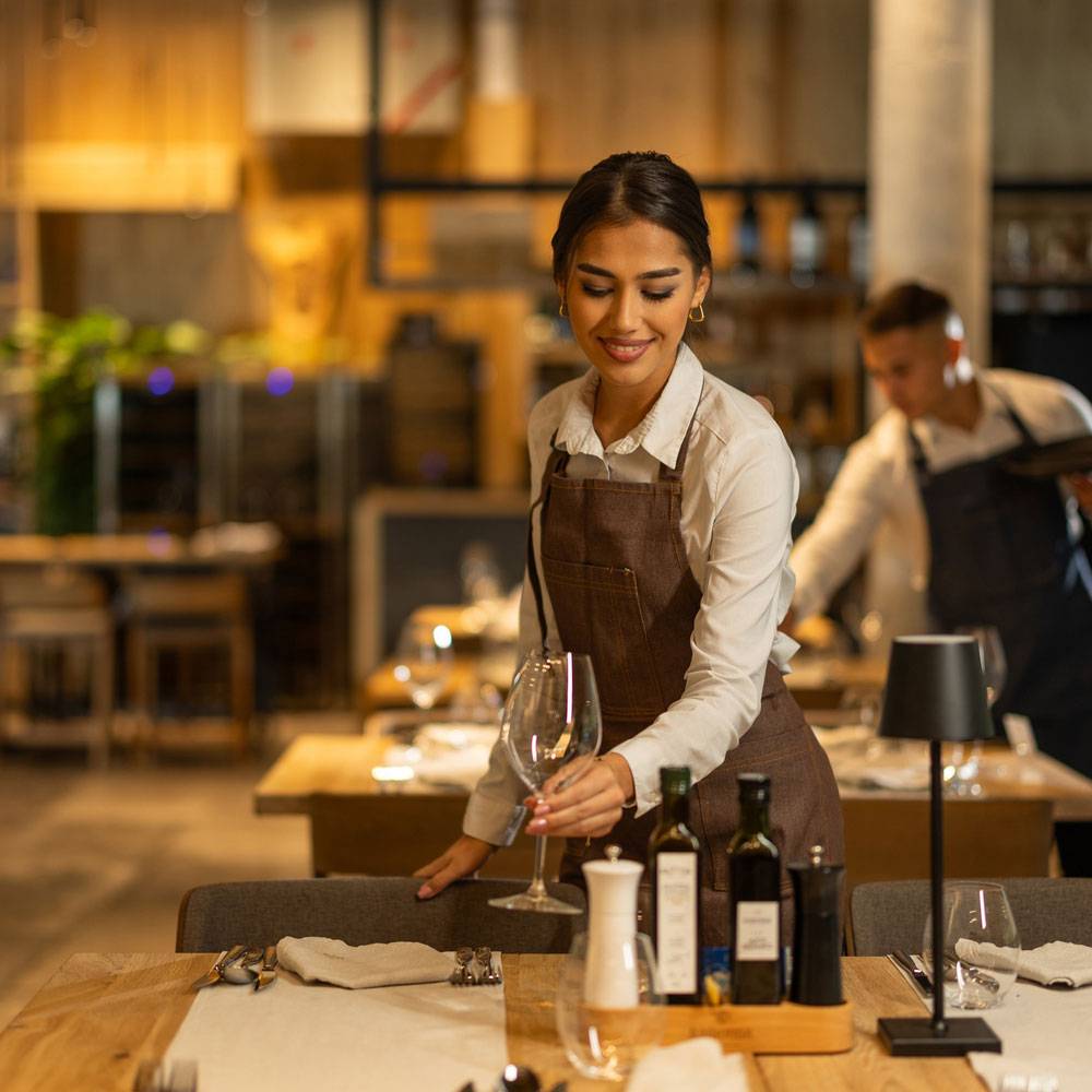 Waitress in brown apron preps dining table with wine glass.