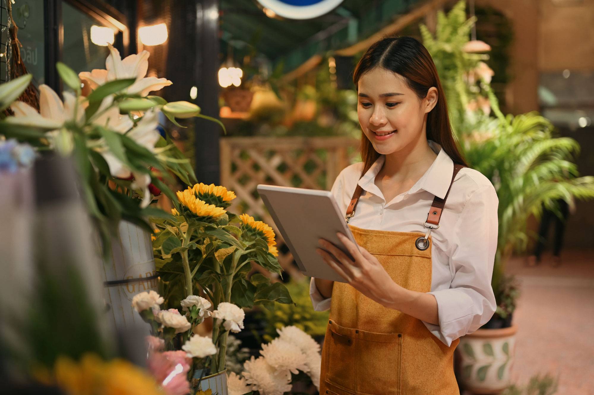 Woman in apron surrounded by flowers in her shop, looking at tablet.