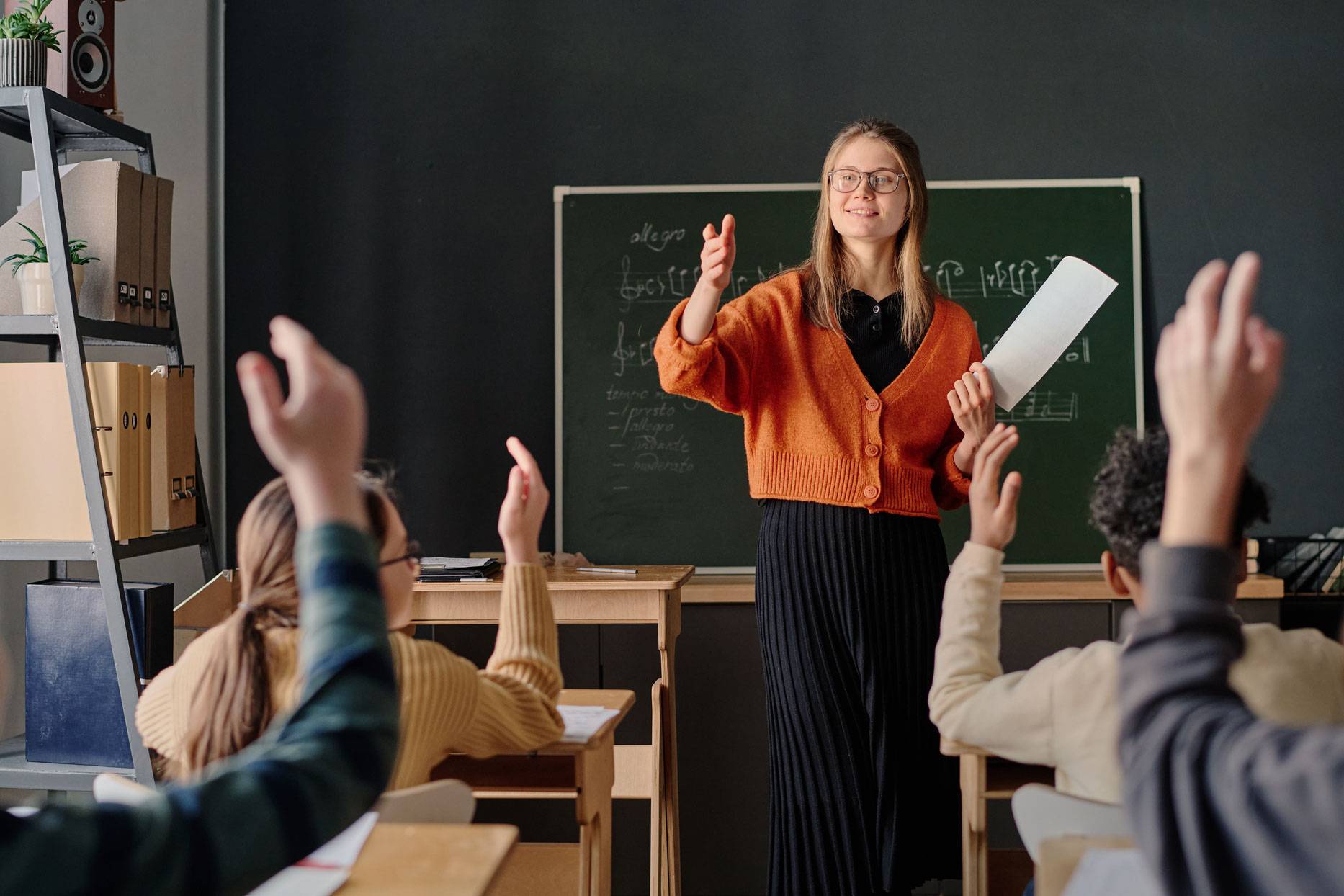 Teacher in front of chalkboard presents to a classroom full of raised hands.