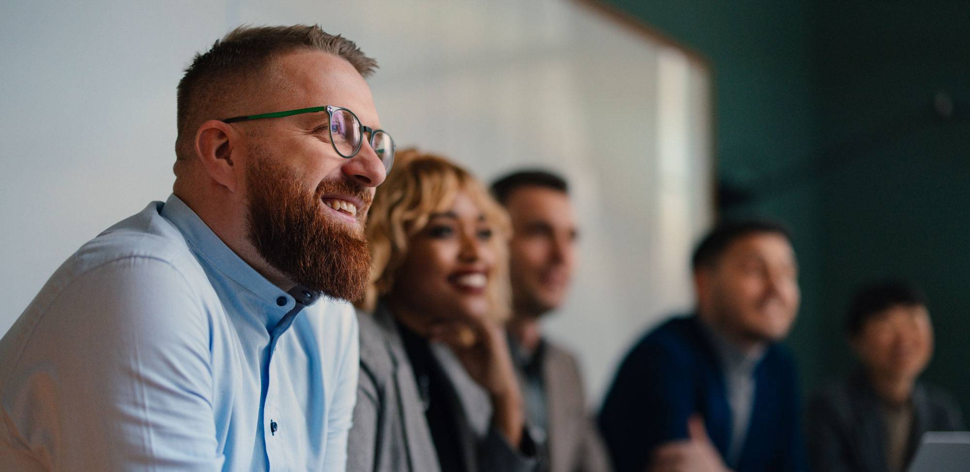 Bearded man in glasses in corporate office setting, with blurred people in the background.