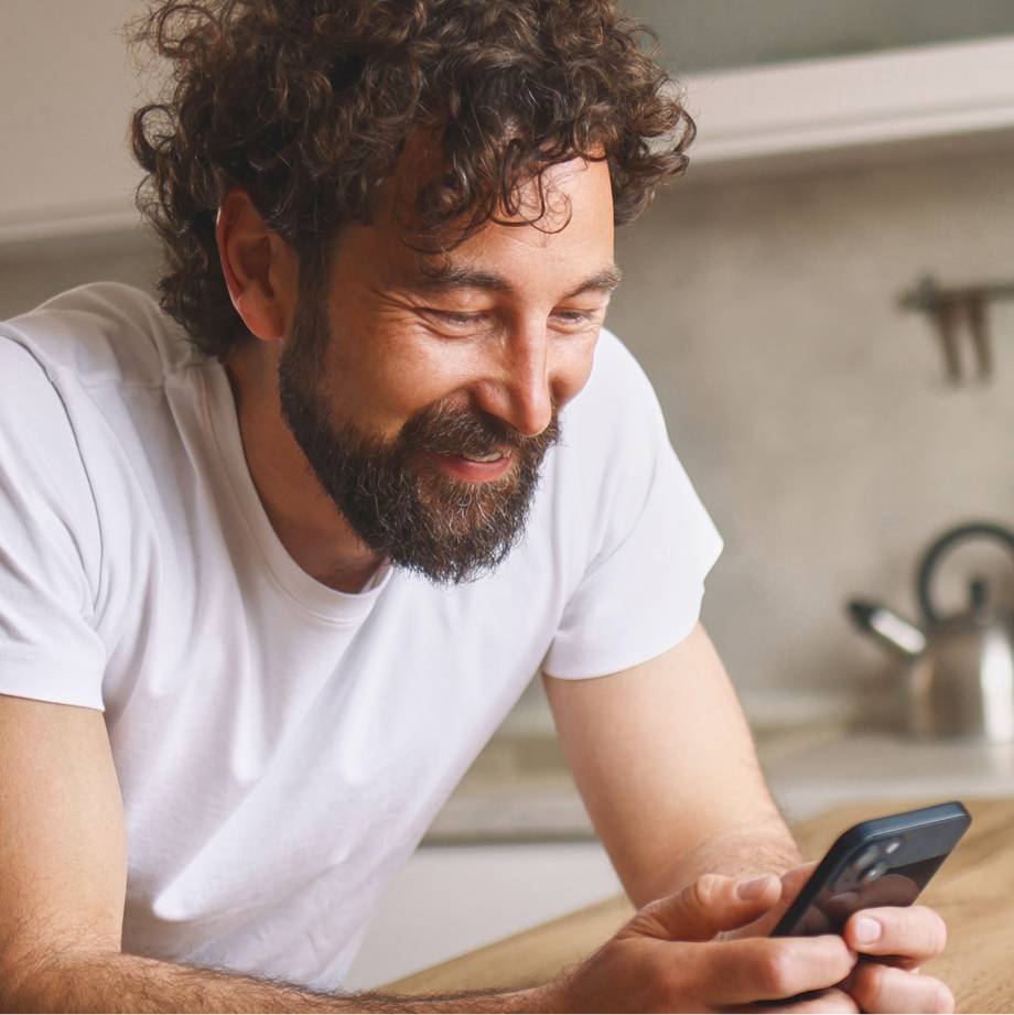 Man in white tee shirt leans on kitchen counter while looking at iPhone.
