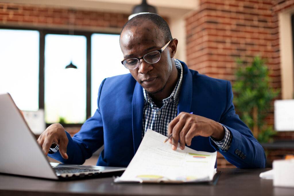 Man in blue blazer sits in front of laptop reviewing paperwork.