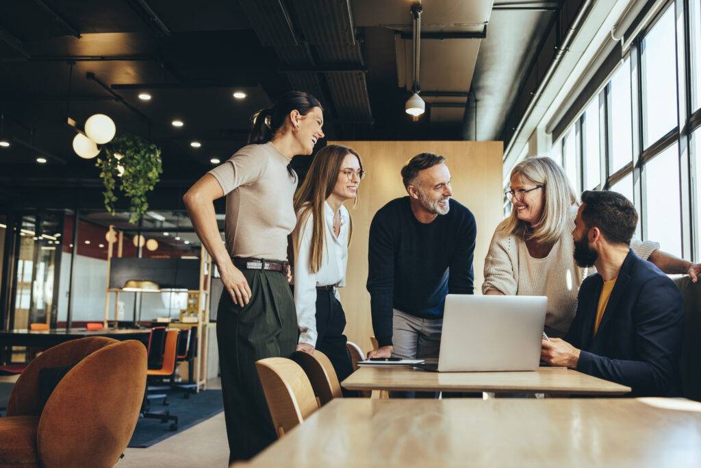 Three women and two men stand around a laptop in a modern corporate office.