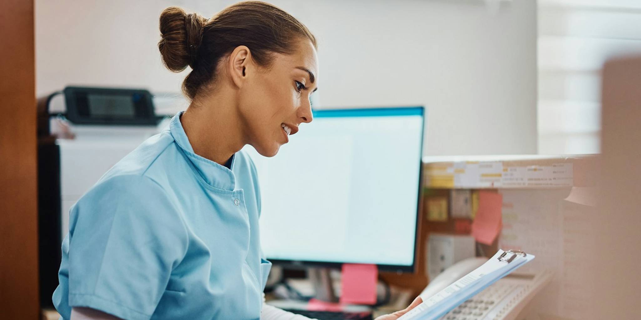 Nurse in scrubs sits at desk reviewing clipboard.