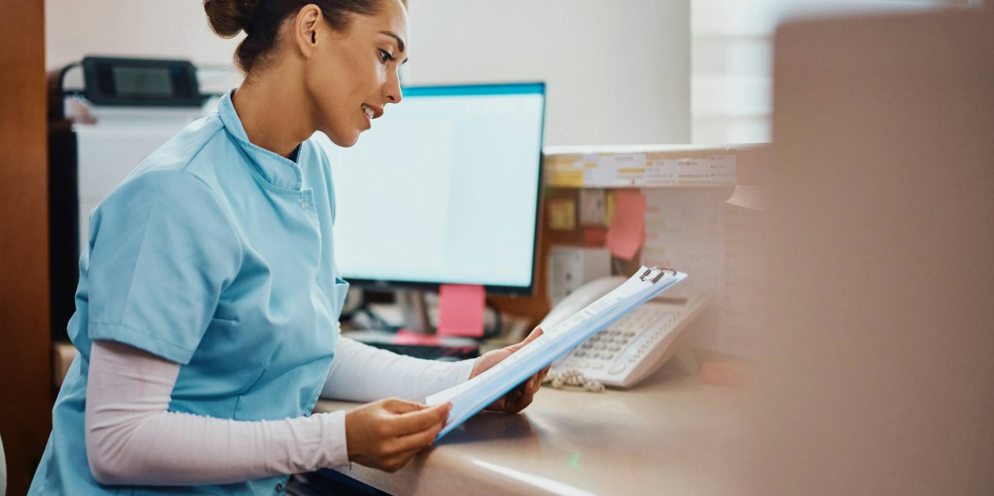 Female nurse in scrubs sits at desk and reviews clipboard.
