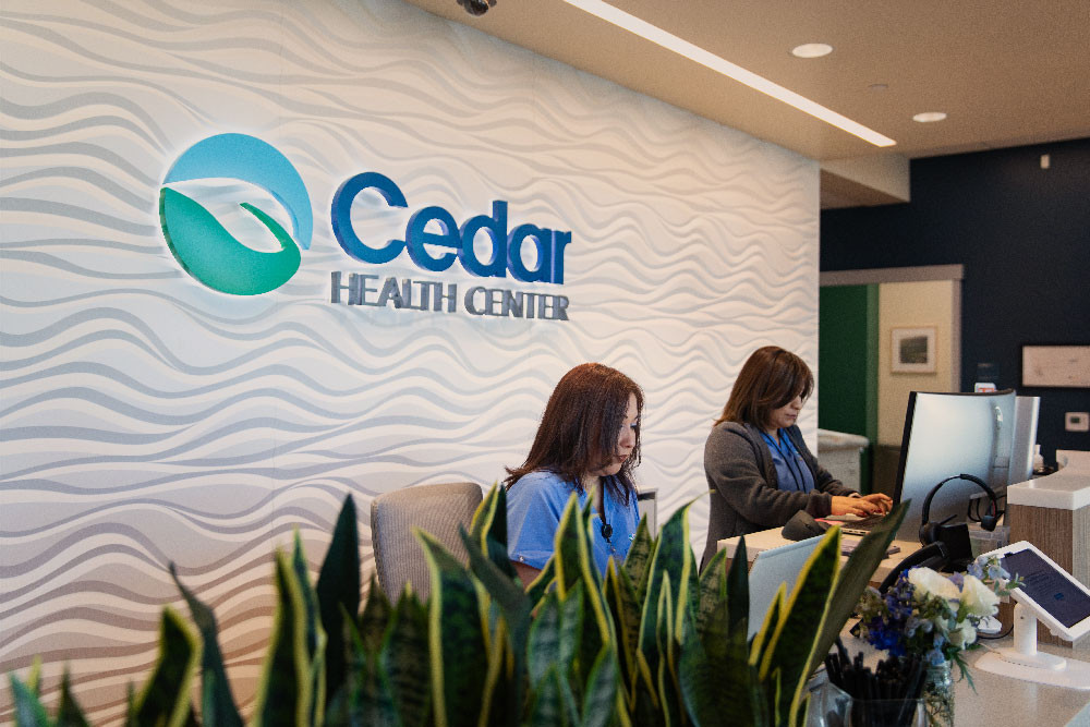 Two receptionists at medical office desk in front of Cedar Health Center logo.