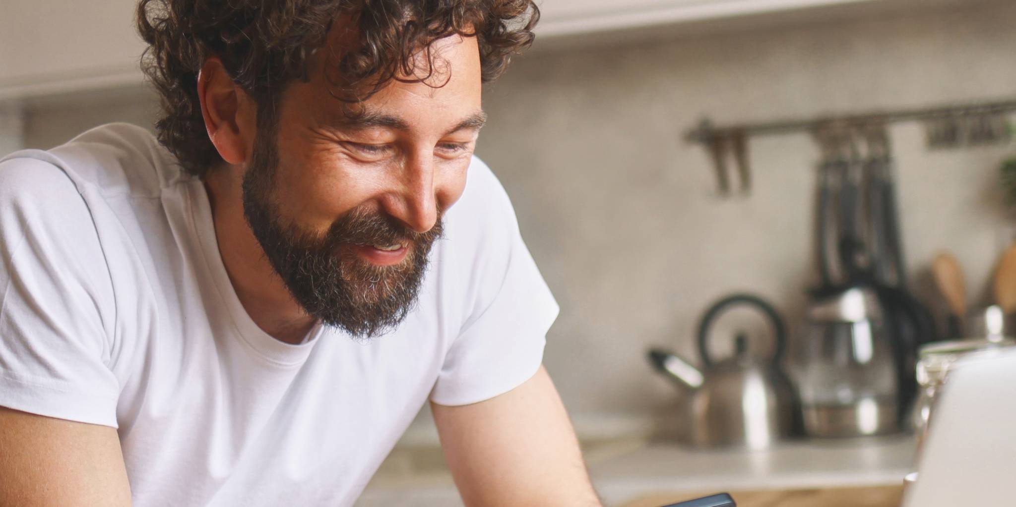 Man in white tee shirt leans on kitchen counter and looks at iPhone