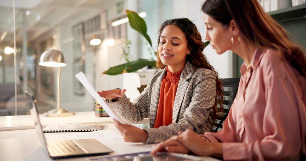 Two women in business attire sit in front of a laptop while looking over printouts.