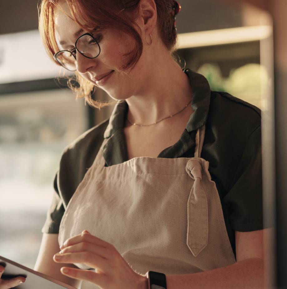 Woman in apron and glasses reviews order on a tablet.