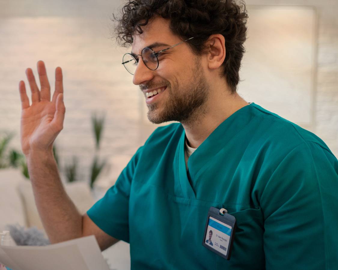 Doctor in green scrubs and glasses raising right hand in greeting.