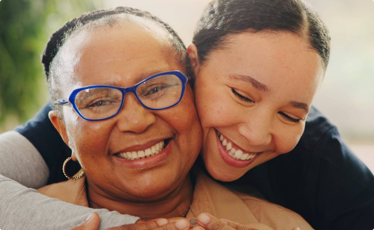 Older woman in blue glasses with middle aged woman hugging her shoulders, both smiling.