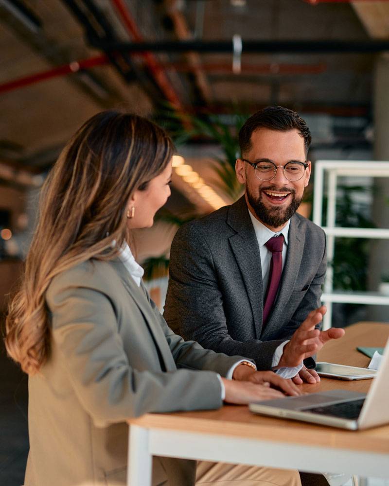 Man and woman in dress attire look at laptop by standing desk.