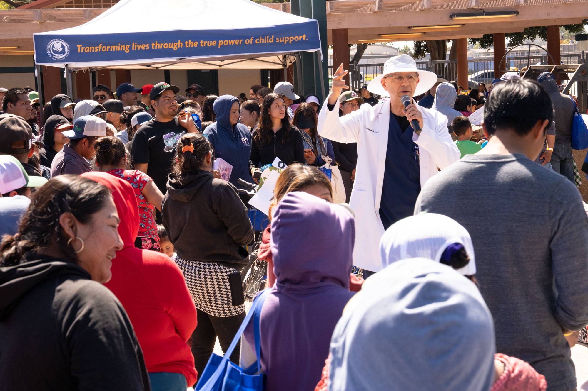 Man in white lab coat and bucket hat speaks to a crowd at a fair.