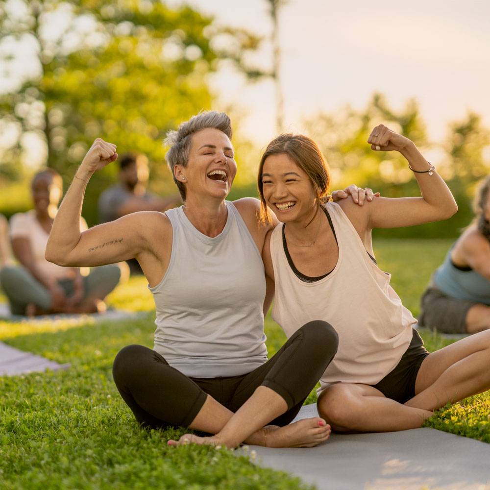 Two women on yoga mat with arms around each other, smiling and flexing their biceps.