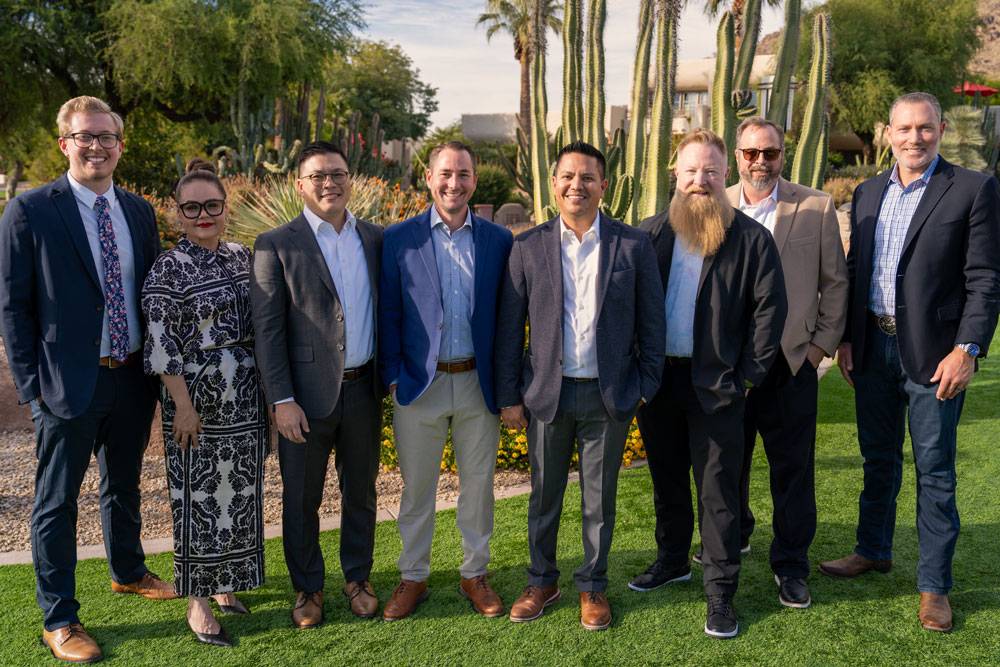 Group of men and women in desert setting, standing in a row in blazers and dresses.