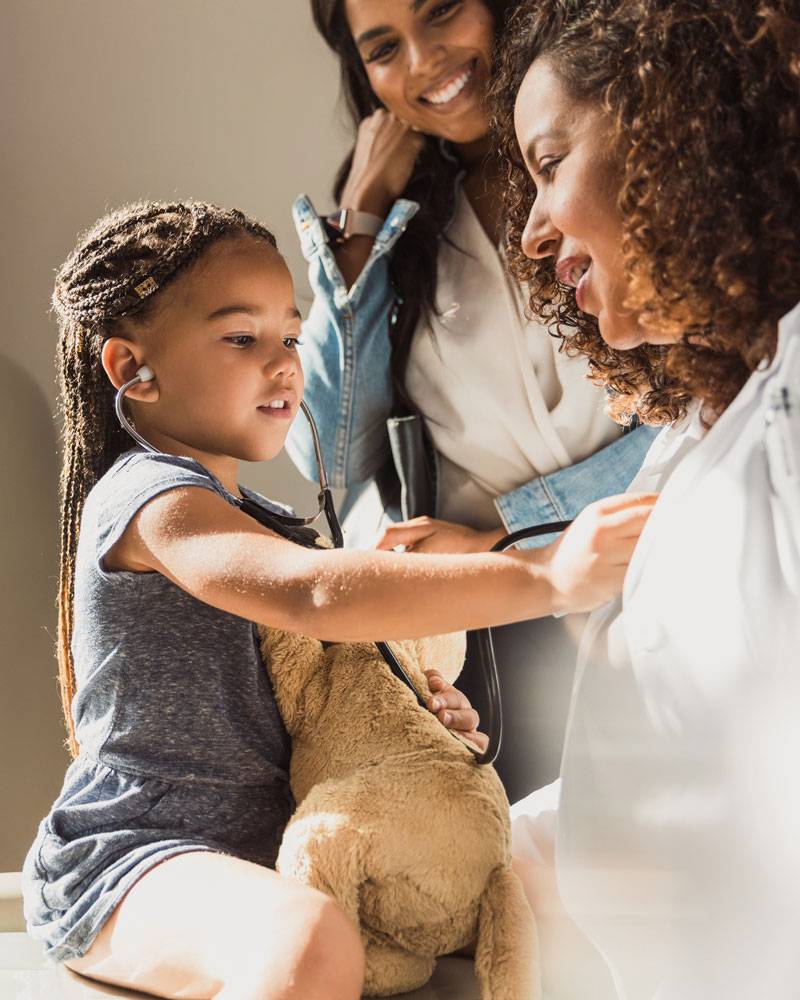 Girl sitting on doctor's lap holding stuffed dog, mom watching in background.
